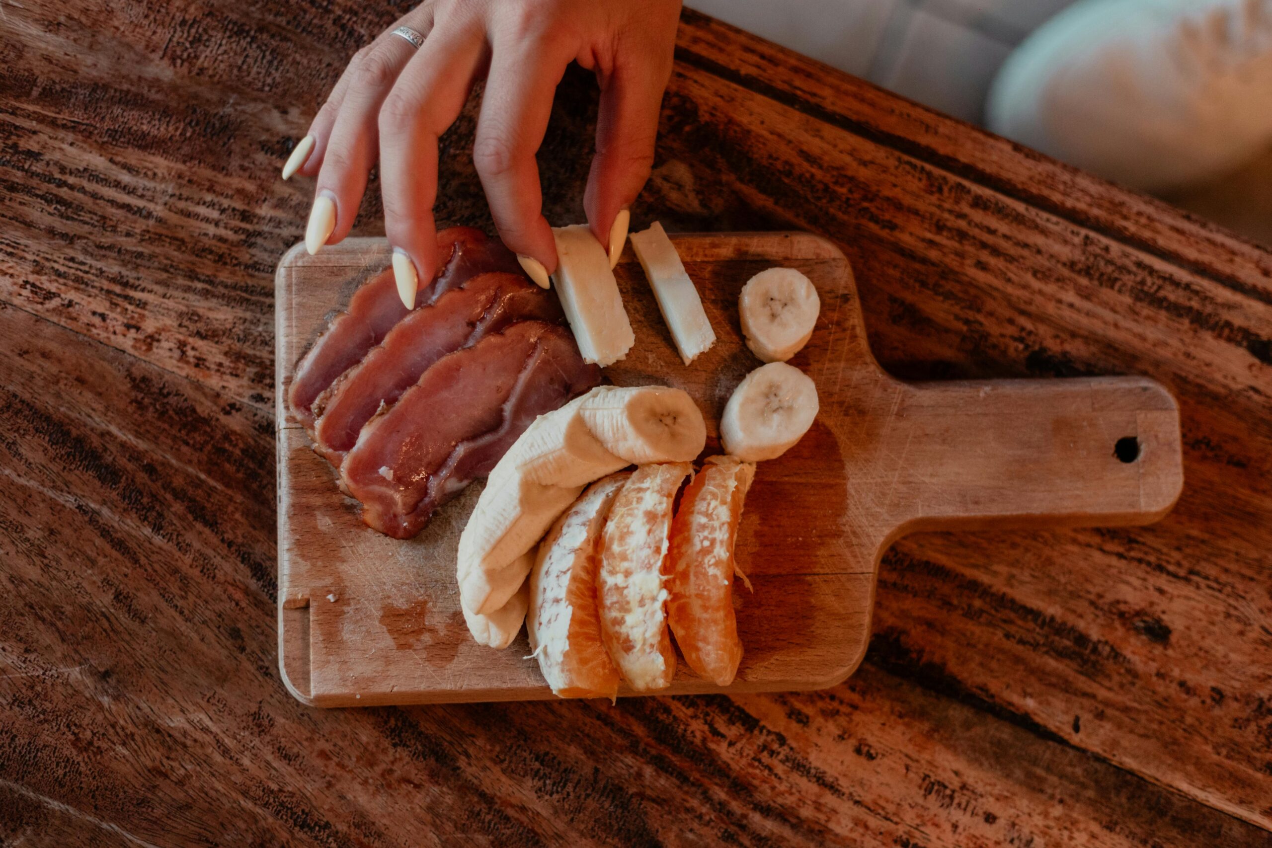 Eine Person bereitet auf einem rustikalen Holzbrett einen gesunden Snack mit Banane, Orangenscheiben und Prosciutto zu.