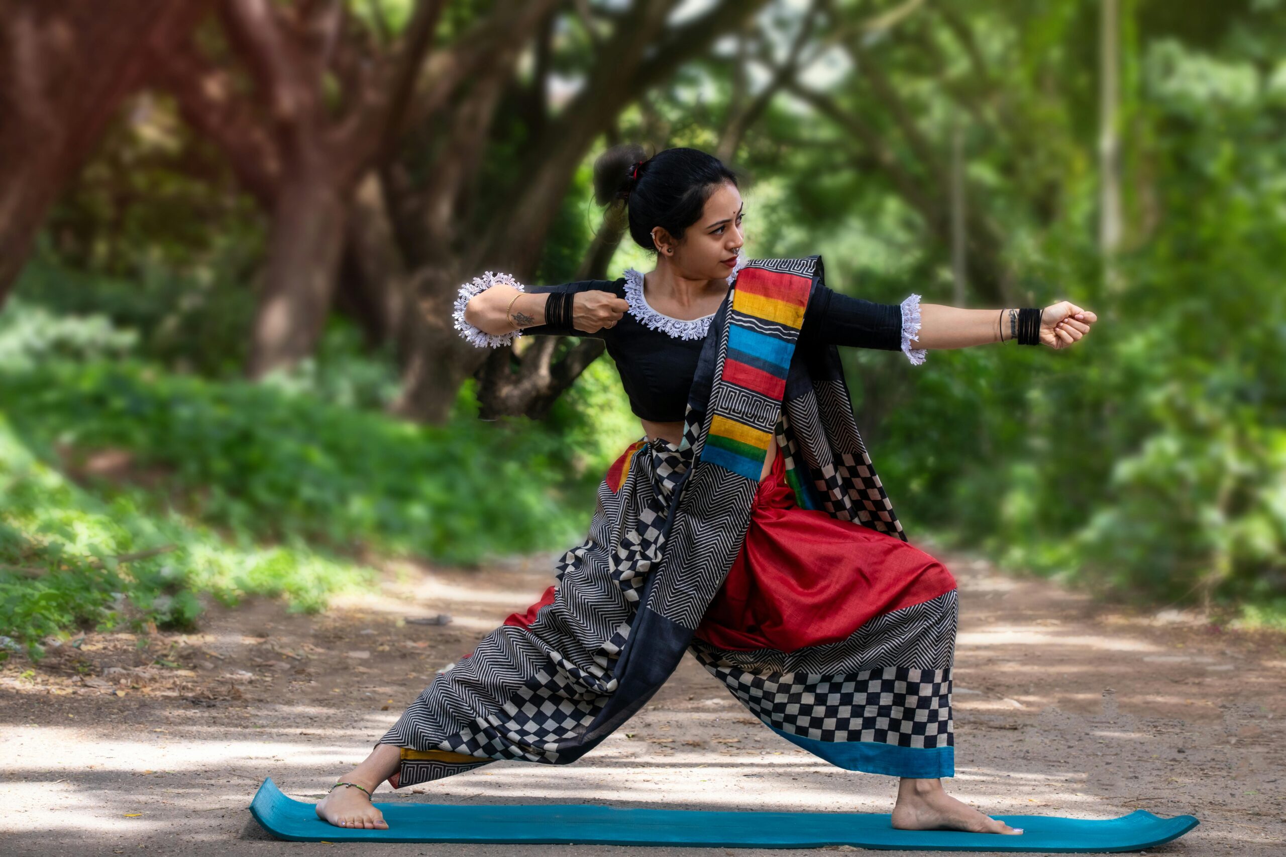Eine Frau praktiziert Yoga in einem farbenfrohen Outfit auf einem Waldweg und verkörpert dabei Achtsamkeit und Tradition.