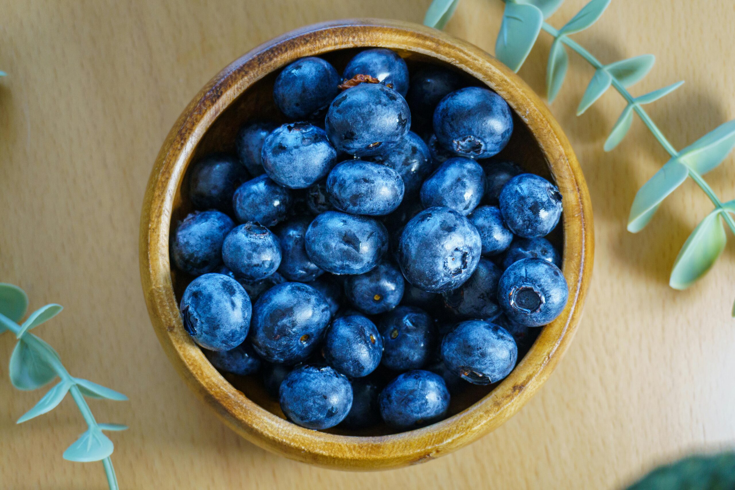 Eine Draufsicht auf frische Blaubeeren in einer Holzschale, umgeben von grünen Blättern, auf einem Holztisch.
