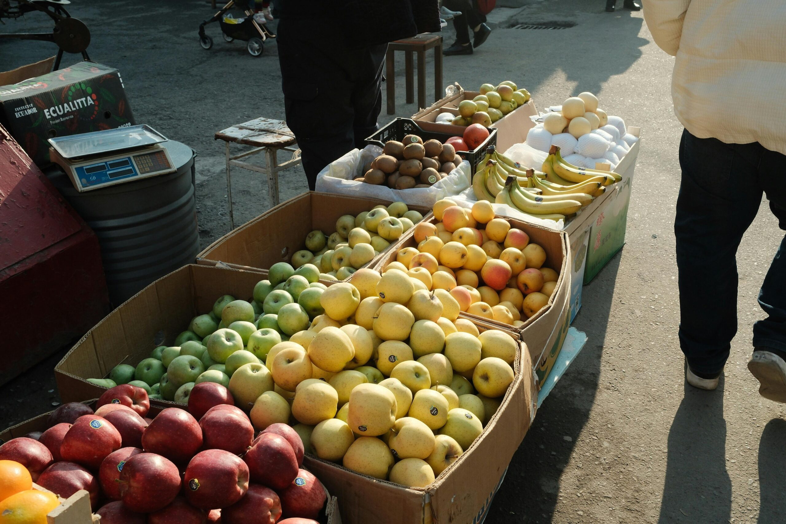 Farbenfroher Obstmarktstand im Freien mit Äpfeln, Kiwis, Bananen und Eiern, die im Sonnenlicht ausgestellt sind.