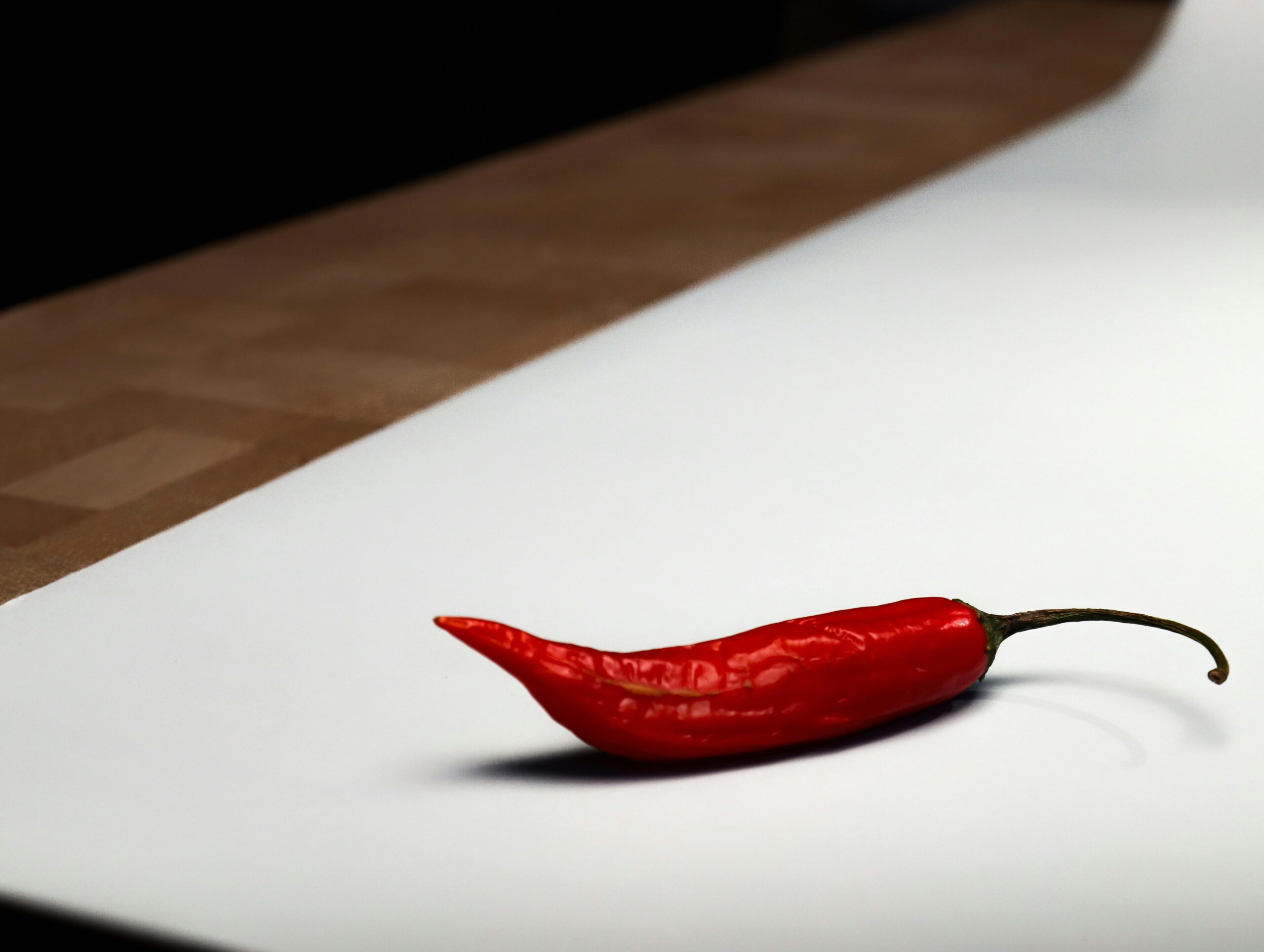 Eine einzelne rote Chilischote liegt auf einer weißen Oberfläche mit sanfter Beleuchtung und minimalistischem Design.
